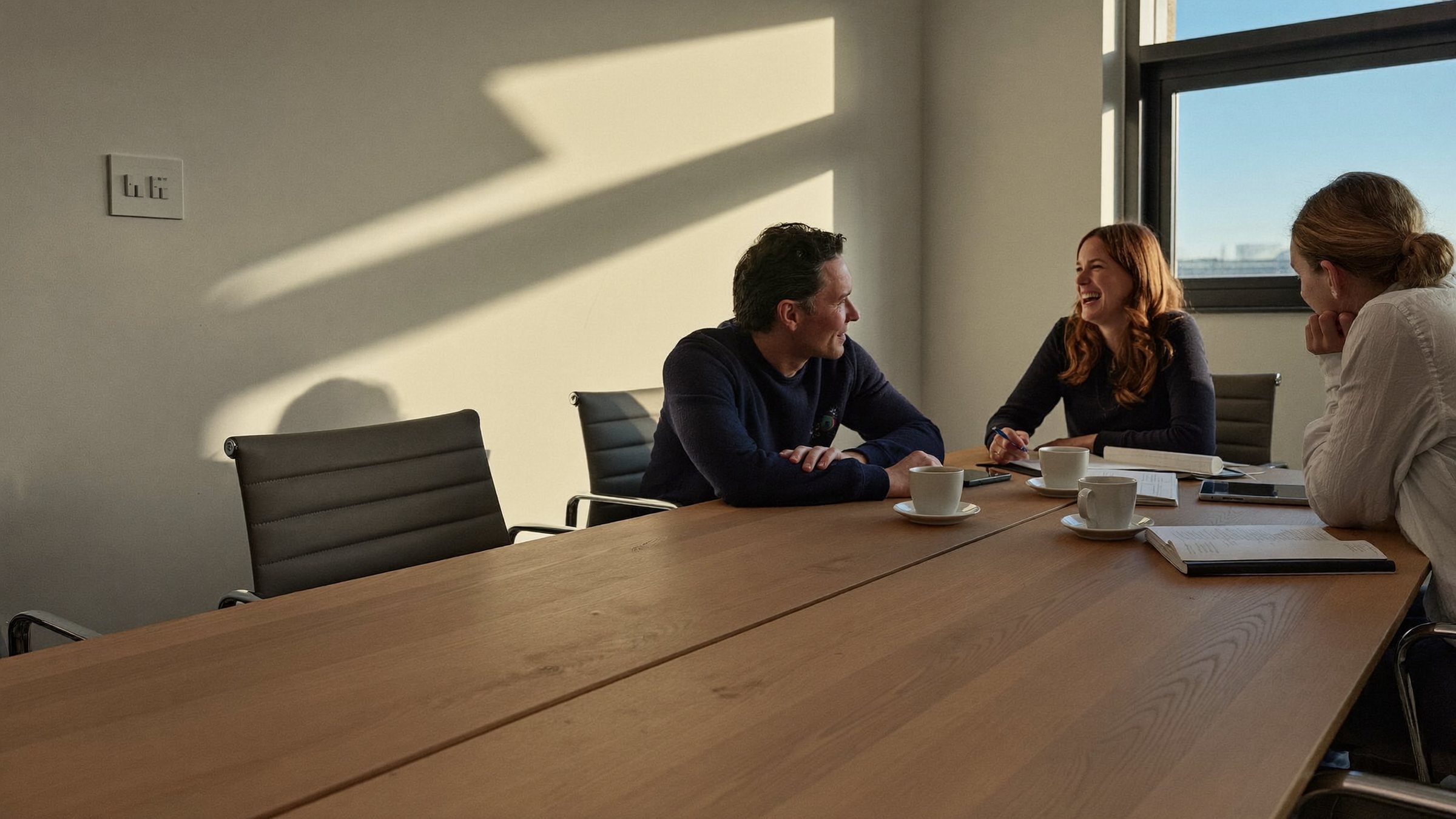 People talking at the far end of a long wooden meeting table in afternoon light