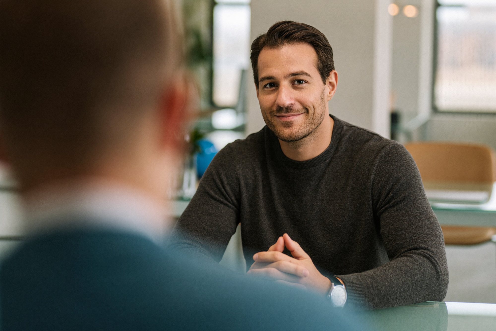 A man smiling across a table while a colleague in the foreground talks