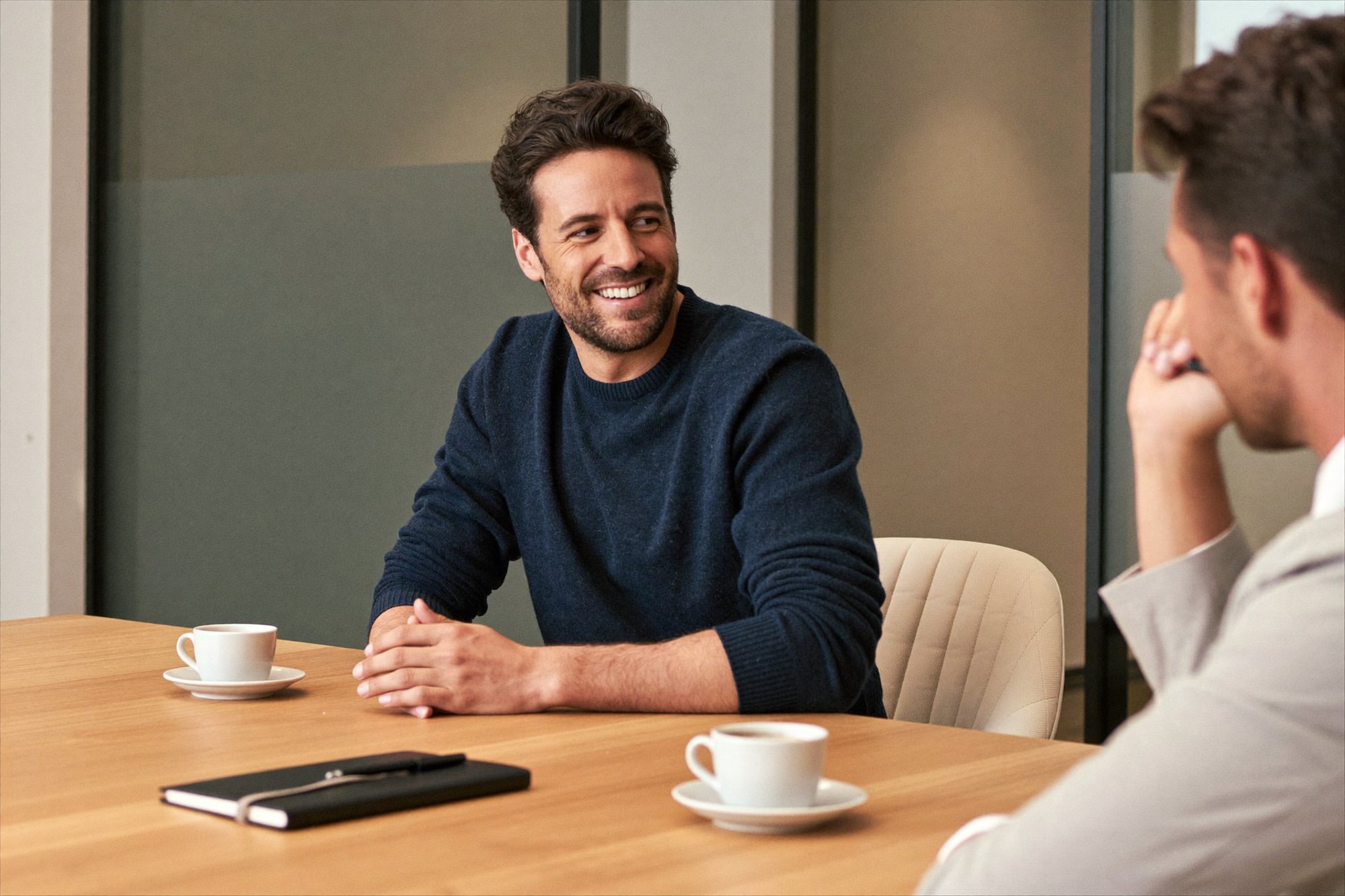 A man in a navy jumper smiling at a meeting table with coffee and a notebook