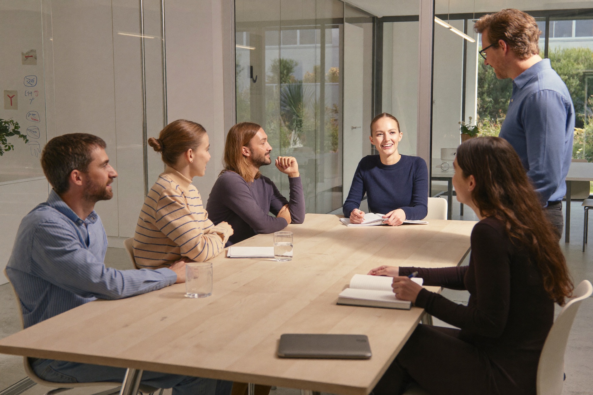 A small team around a meeting table, one standing to talk while the others listen
