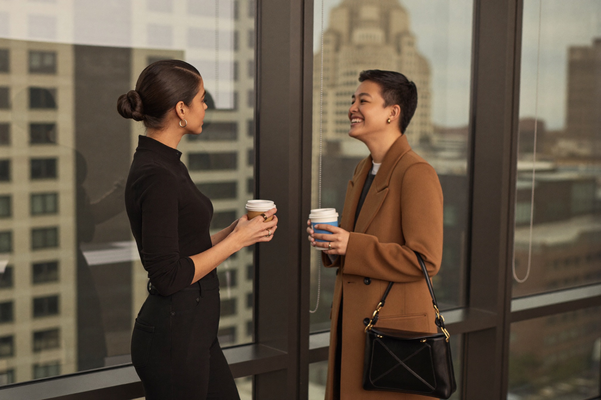 Two women holding coffee cups and talking beside a tall window
